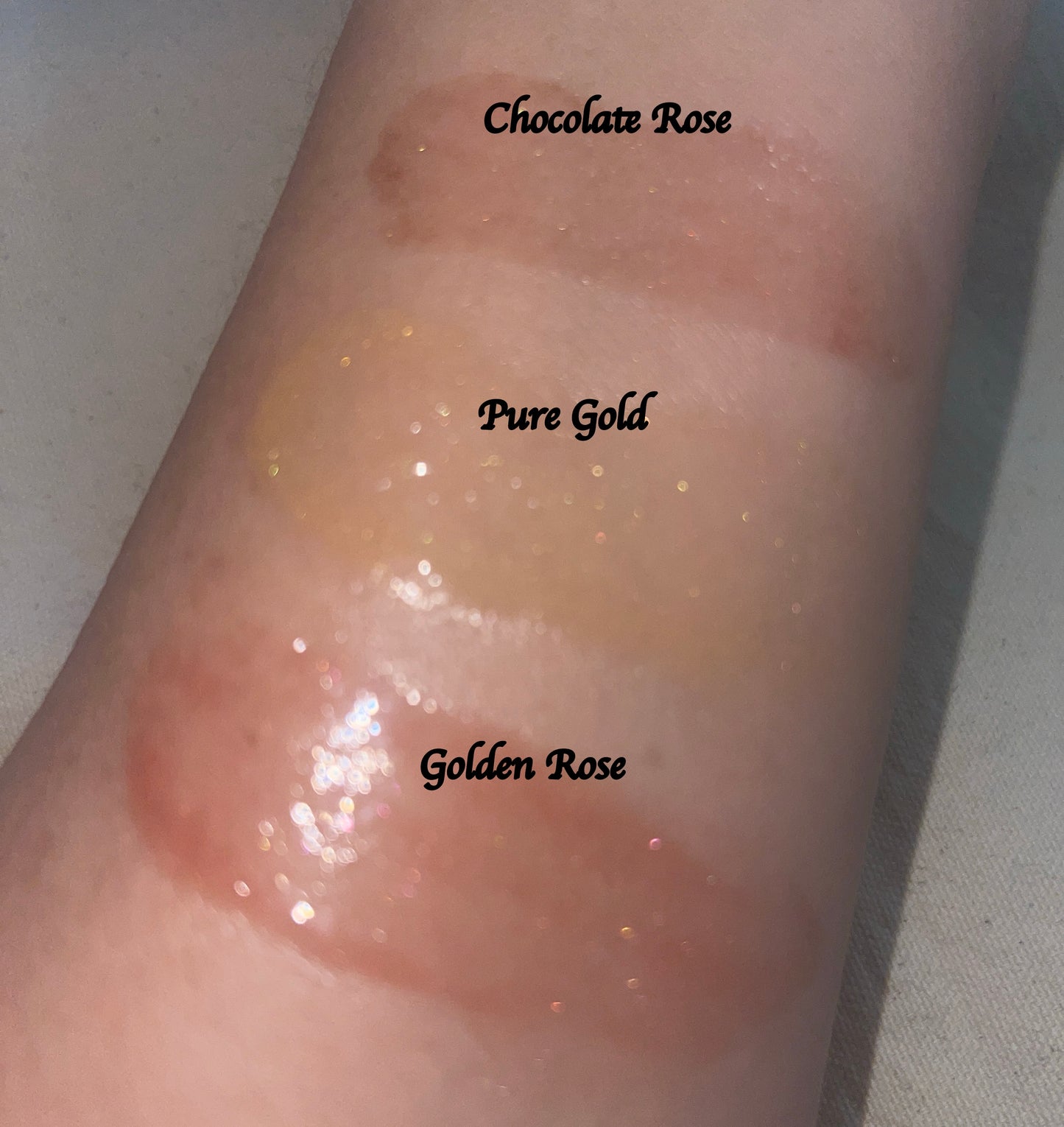 Three swatch shades of glittery face balm gloss applied to a person's arm with labels 'Chocolate Rose', 'Pure Gold', and 'Golden Rose'.